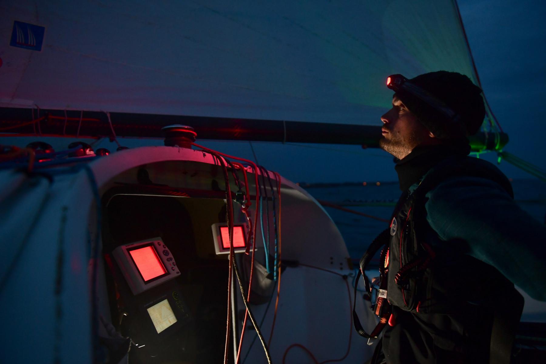 Skipper de nuit avec lampe frontale observant ses instruments de navigation rouges sur un voilier Mini 6.50.