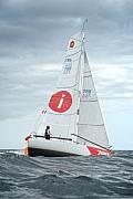 Voilier de course Mini 6.50 avec voiles blanches et logo rouge naviguant sur mer houleuse sous ciel gris.