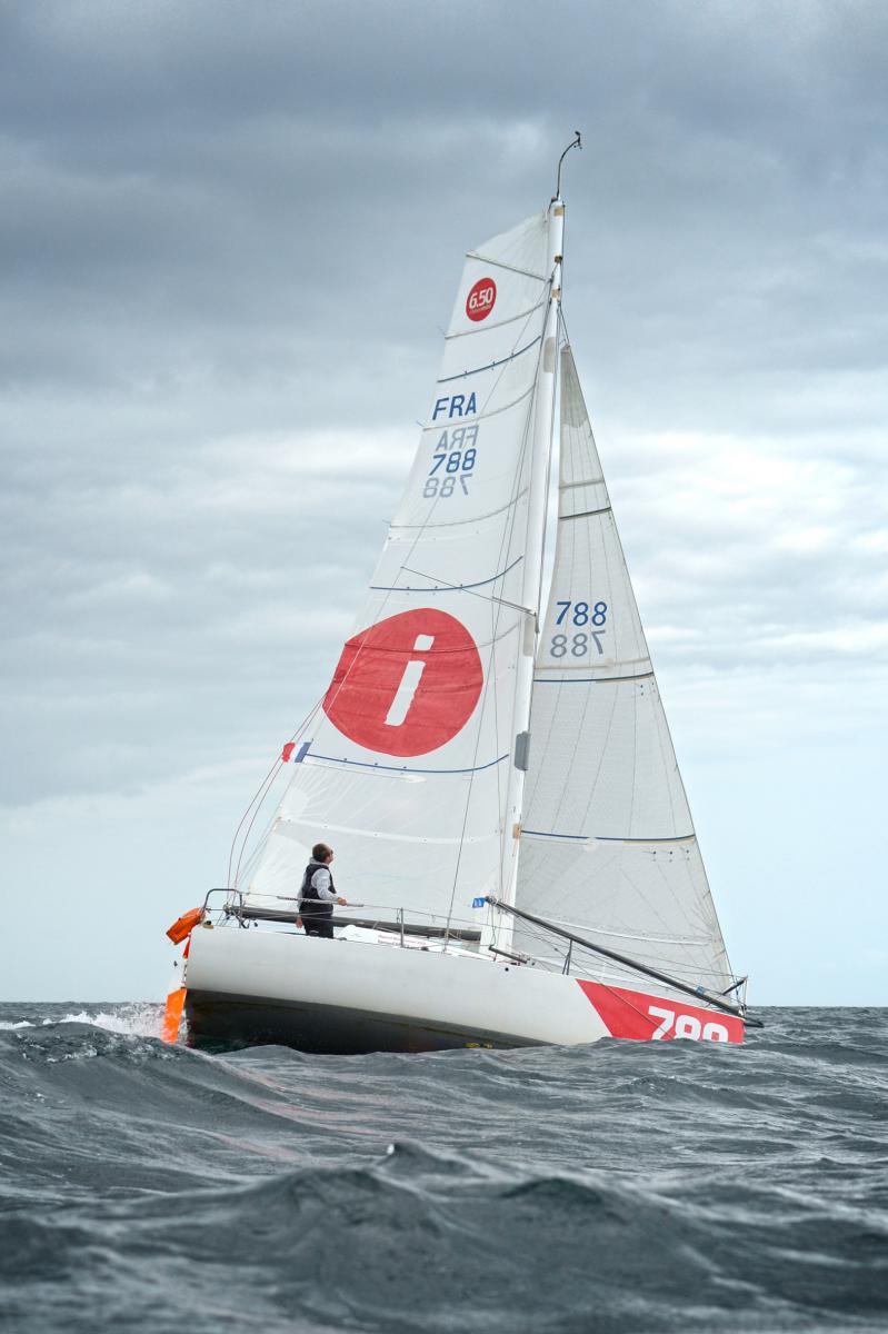 Voilier de course Mini 6.50 avec voiles blanches et logo rouge naviguant sur mer houleuse sous ciel gris.