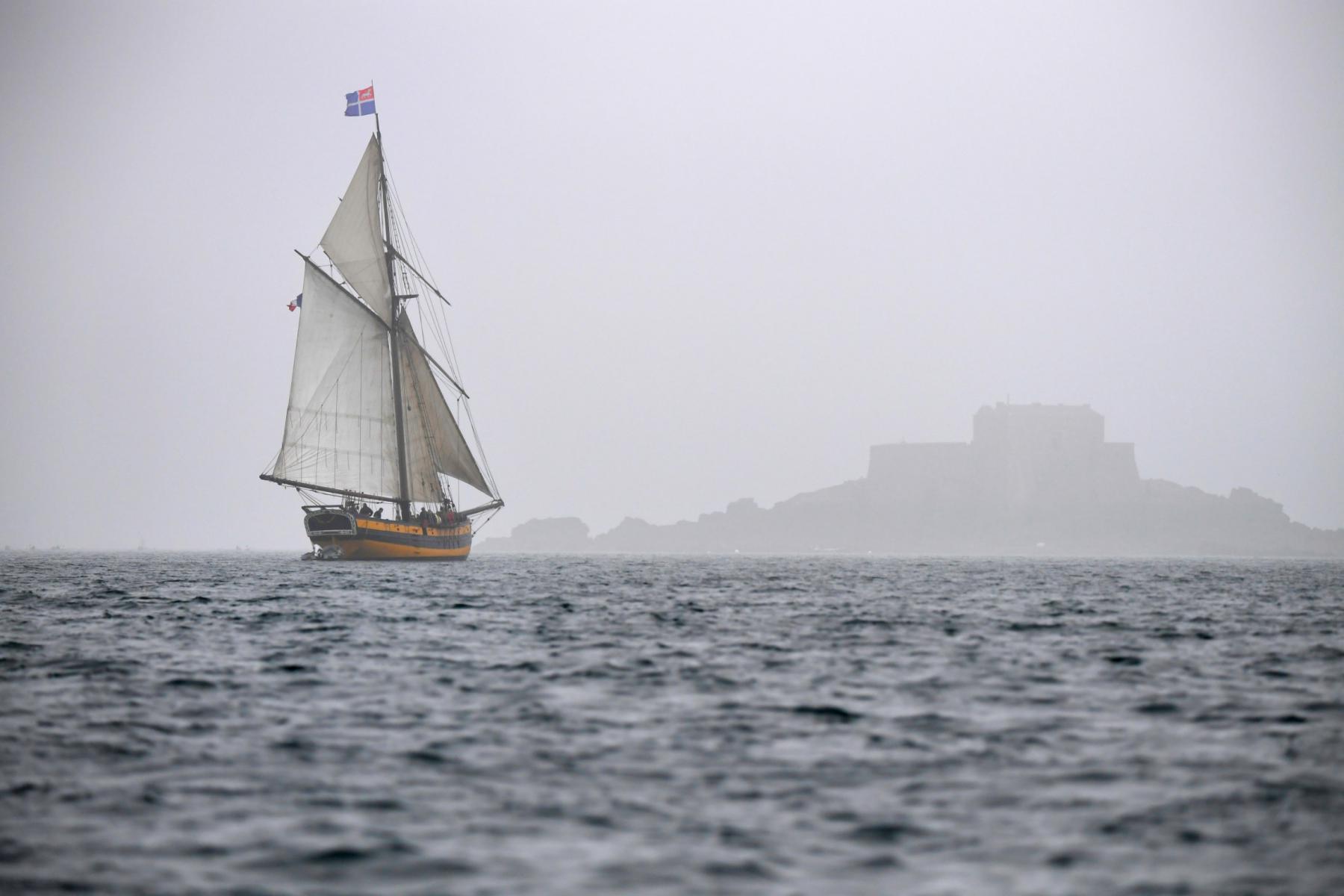 Cotre corsaire Le Renard naviguant toutes voiles dehors dans le brouillard devant la silhouette de Saint-Malo.