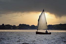 Silhouette de canot à voile sur mer scintillante au coucher du soleil avec nuages sombres et lumière dorée.