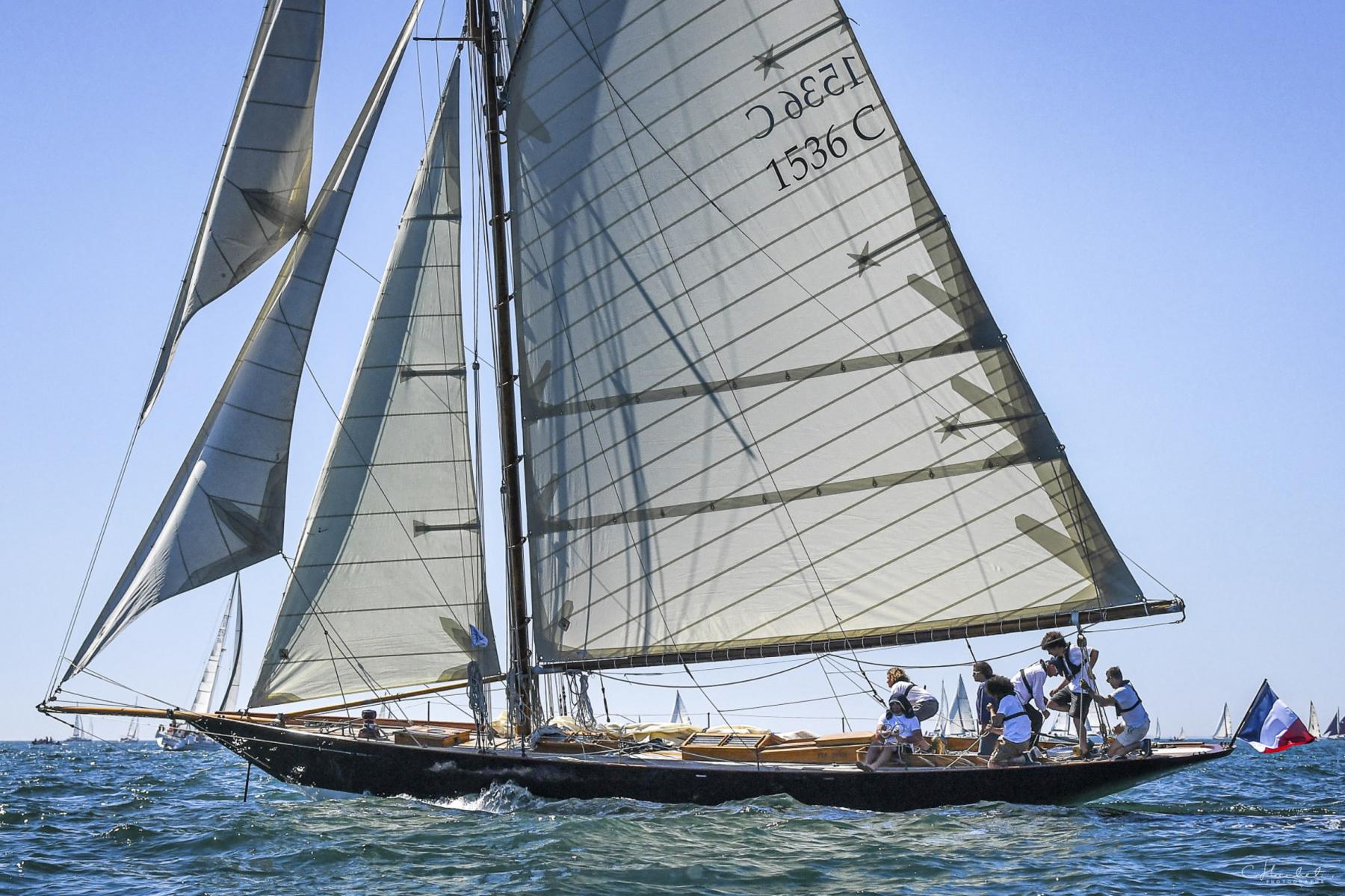 Voilier classique Pen Duick I avec voiles blanches déployées sur mer calme sous un ciel bleu azur