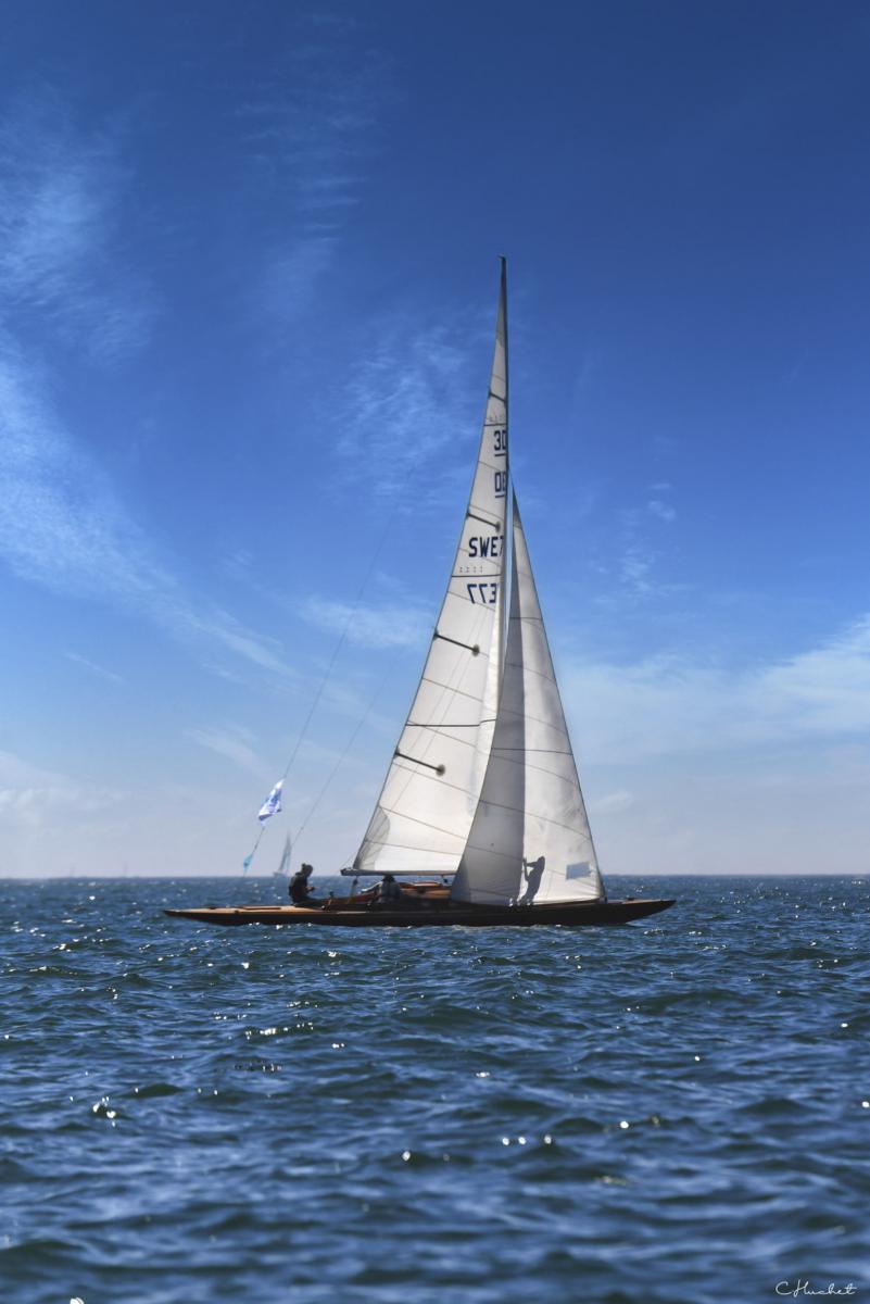 Voilier en bois à coque sombre avec grande voile blanche sur mer bleue scintillante sous ciel azur ensoleillé.