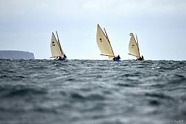 Trois vieux gréements aux voiles crème naviguent de front sur une mer formée devant les falaises du Cap Fréhel.