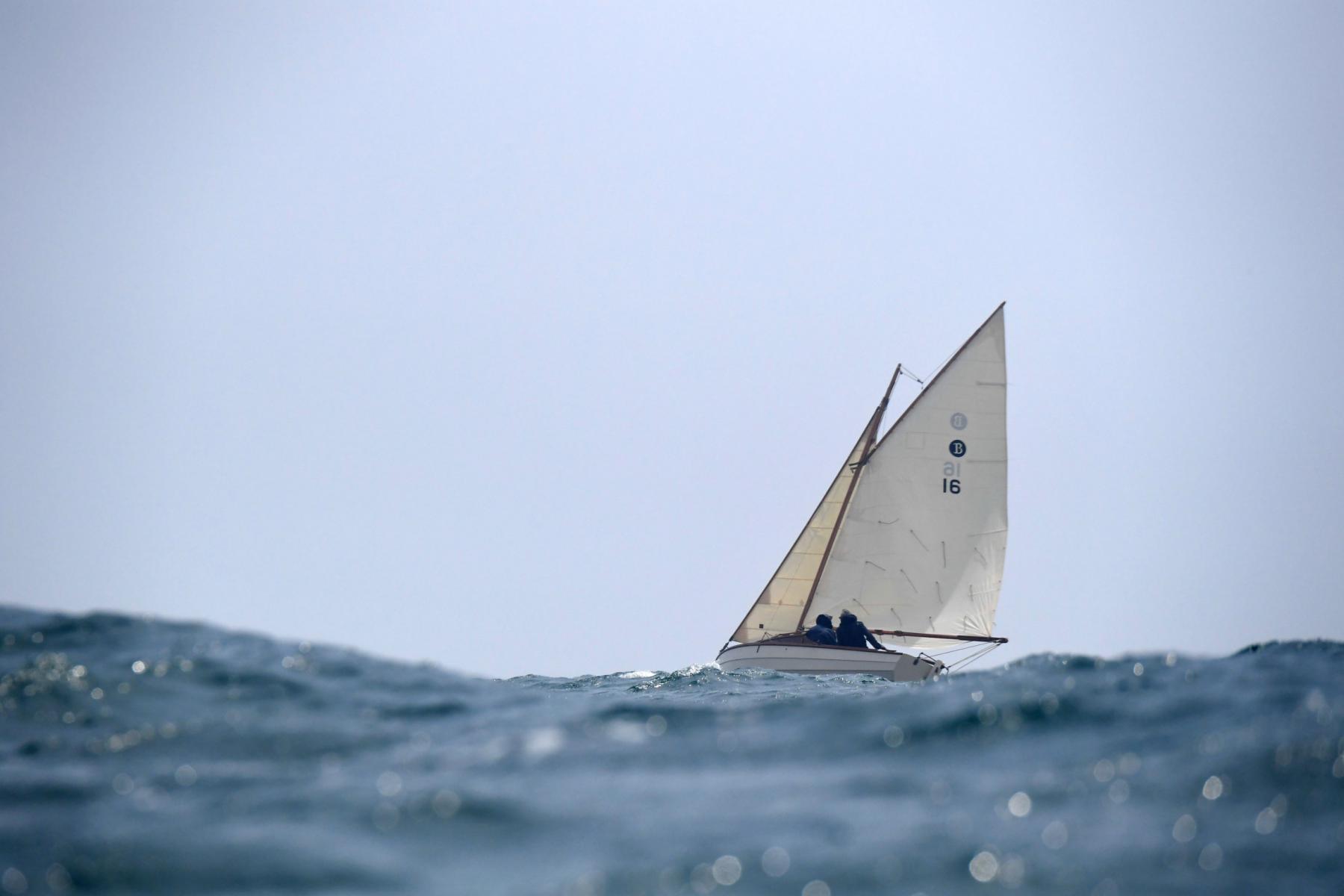 Vieux gréement à voiles blanches naviguant sur une mer agitée sous un ciel clair. Focus sur la gîte du bateau.