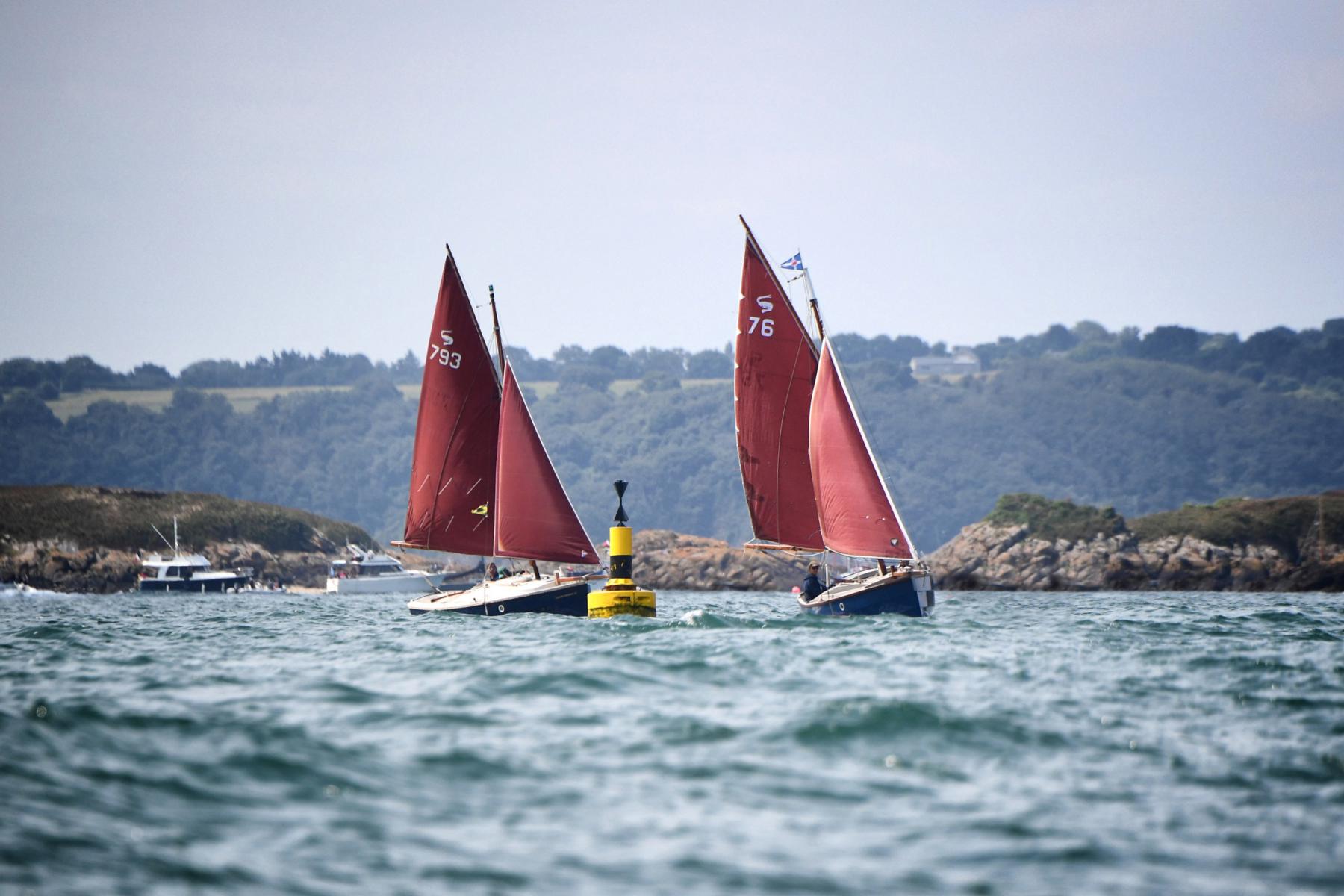 Deux voiliers anciens à voiles briques naviguent près d’une balise jaune devant les îles Ebihens en Bretagne.