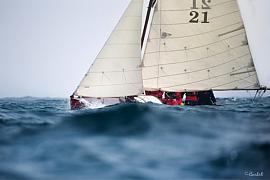 Vieux gréement à voiles crème et coque rouge sur une mer agitée, équipage visible en mouvement.