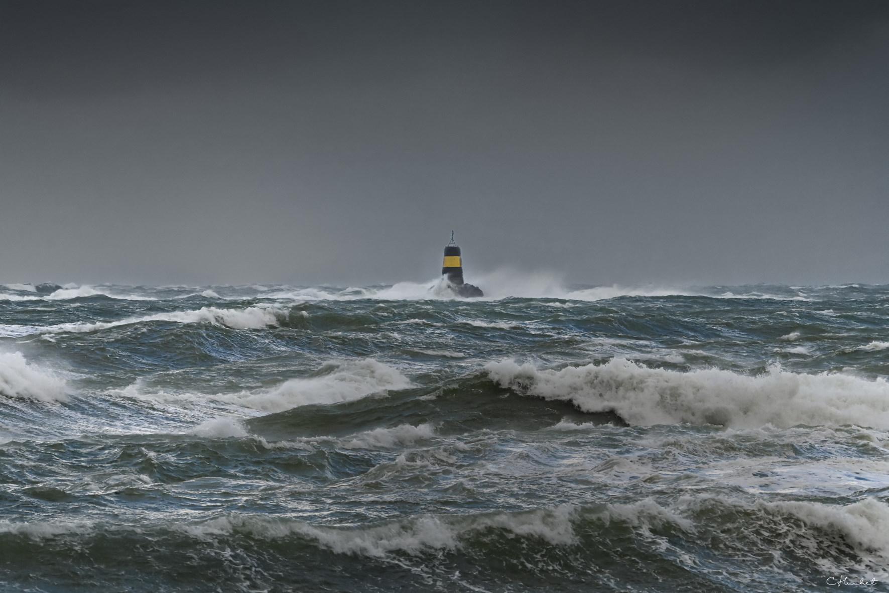 Vagues puissantes et écume blanche entourant la balise jaune et noire Nerput à Saint-Lunaire sous un ciel de tempête sombre.