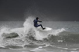Surfeur en combinaison noire en plein virage sur une vague avec projections d’eau sur fond de ciel gris breton.