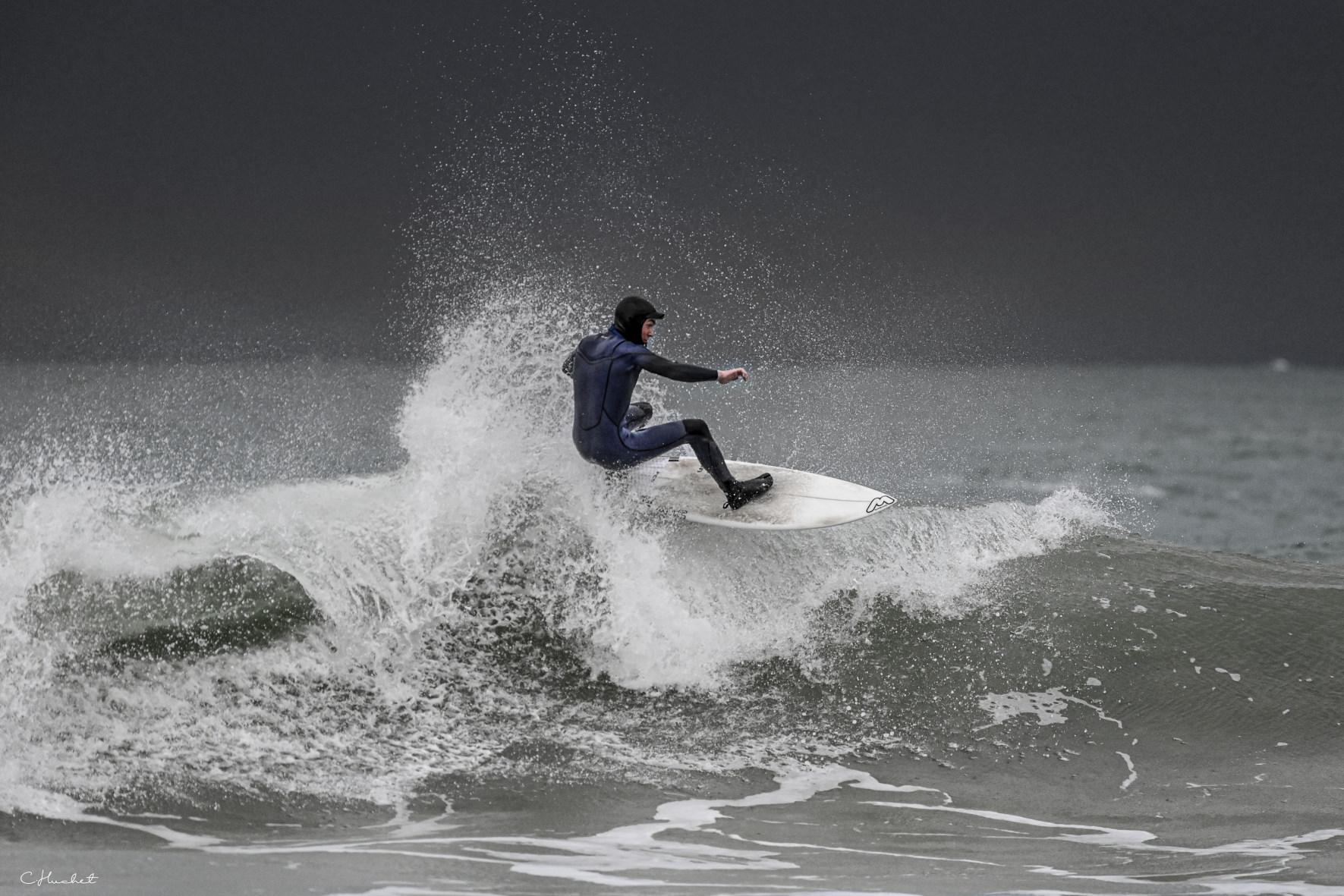Surfeur en combinaison noire en plein virage sur une vague avec projections d’eau sur fond de ciel gris breton.