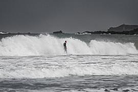 Surfeur en combinaison noire glissant sur une déferlante d’écume blanche sous un ciel gris à la plage de Longchamps.