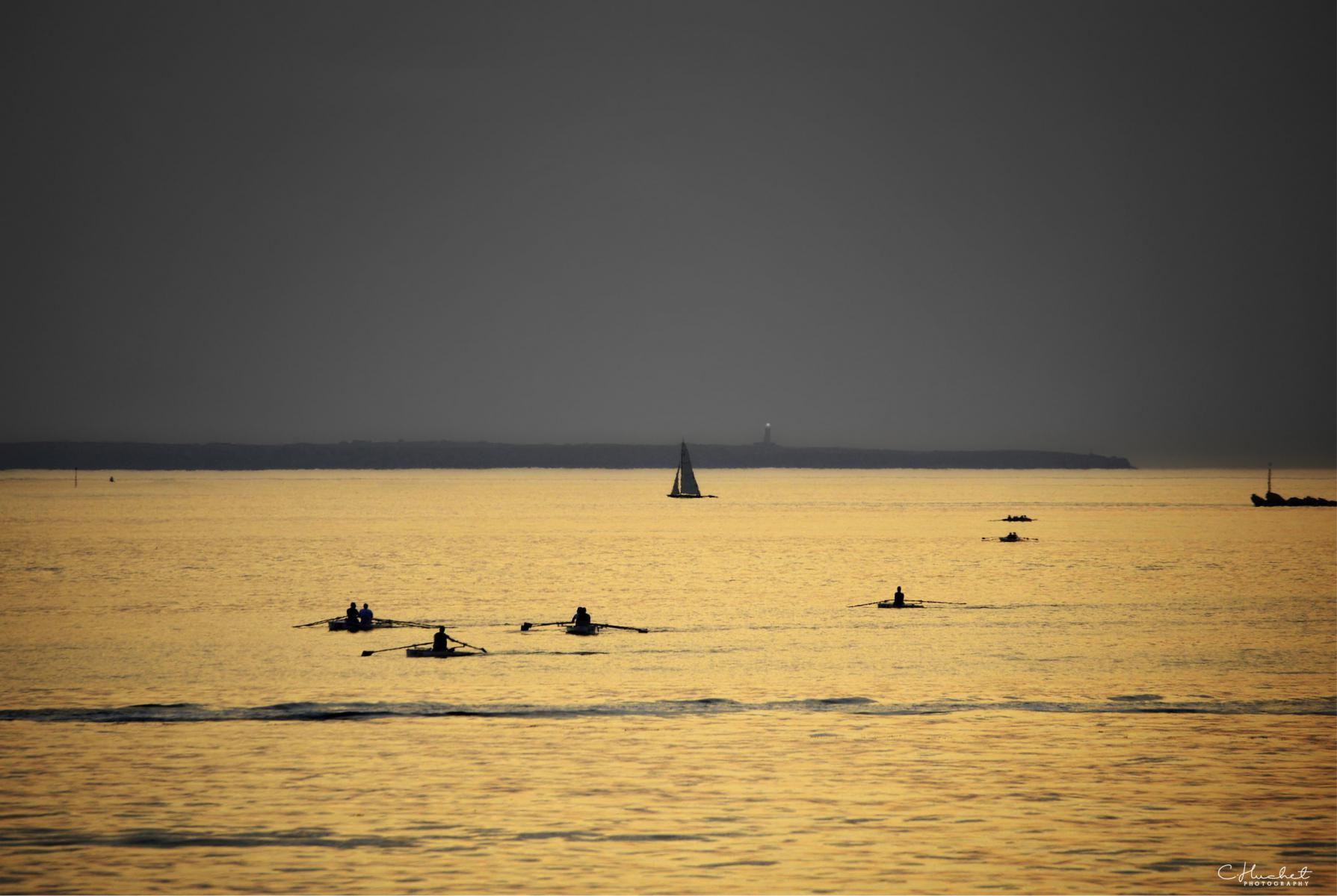 Silhouettes de rameurs en aviron sur une mer dorée au crépuscule face à la côte de Saint-Malo en Ille-et-Vilaine.
