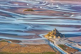 Vue aérienne du Mont Saint-Michel à marée basse montrant les méandres d’eau et l’îlot Tombelaine sous une lumière rasante