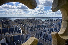 Toits d’ardoise de Saint-Malo vus depuis une ouverture en pierre sculptée sous un ciel nuageux et la mer au loin.