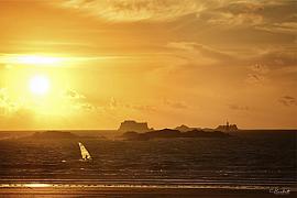Planche à voile naviguant sur mer agitée devant Saint-Malo sous un ciel orange intense de soleil couchant.