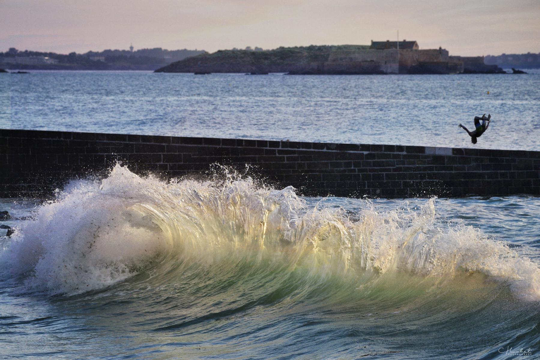 Silhouette réalisant un salto sur une digue devant une vague puissante, éclairage naturel, fort de Saint-Malo au second plan.