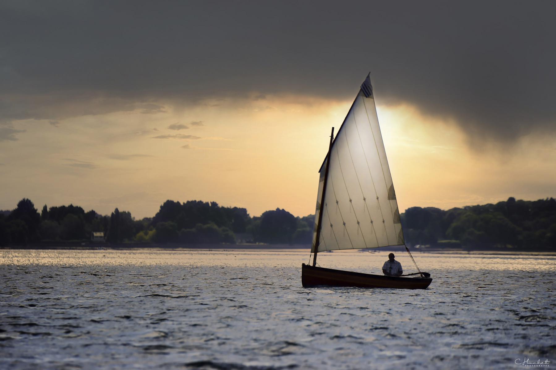 Voilier traditionnel à une voile naviguant au crépuscule sur l’eau scintillante sous un ciel de tempête orangé.