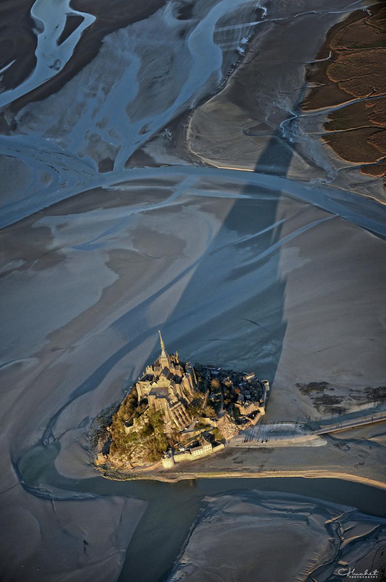 Vue aérienne verticale du Mont Saint-Michel et sa longue ombre portée sur le sable de la baie à marée basse.