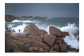Éclats d’écume blanche sur rochers de granit rose. Deux silhouettes observent la mer déchaînée sous un ciel sombre et menaçant.