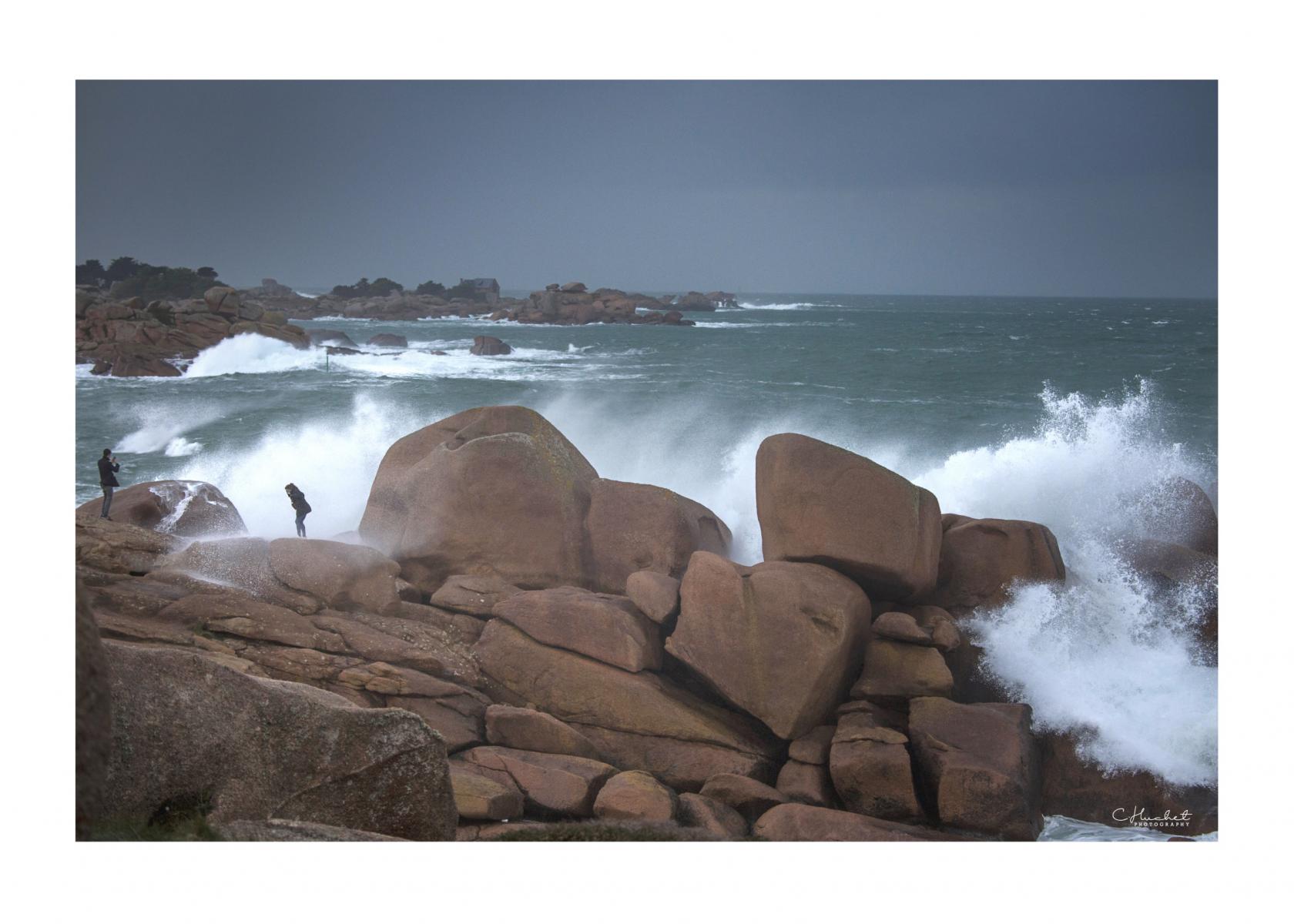 Éclats d’écume blanche sur rochers de granit rose. Deux silhouettes observent la mer déchaînée sous un ciel sombre et menaçant.