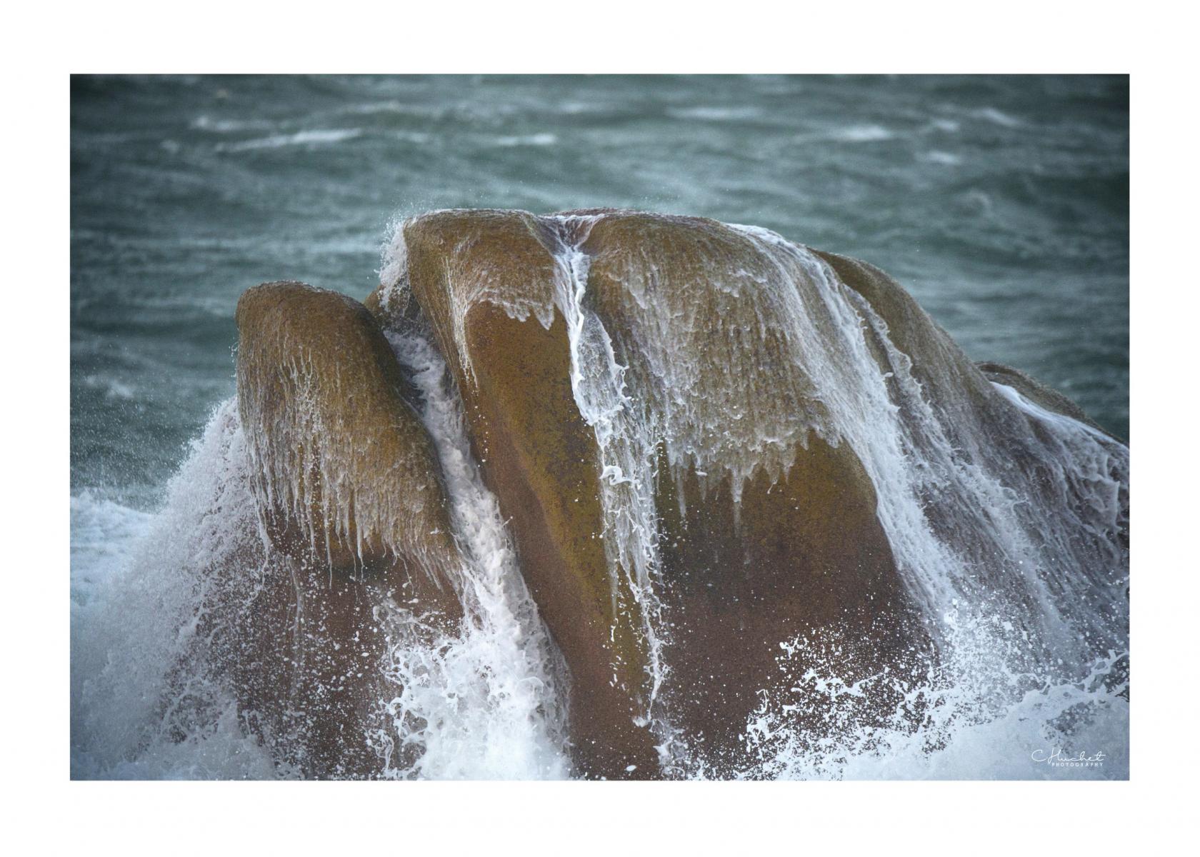Rocher anthropomorphe frappé par l’écume en mer d’Iroise. Eau ruisselante sur le granit sous une lumière naturelle diffuse.
