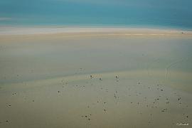 Immense étendue de sable blond parsemée de roches à marée basse avec quelques silhouettes humaines sous une mer turquoise.