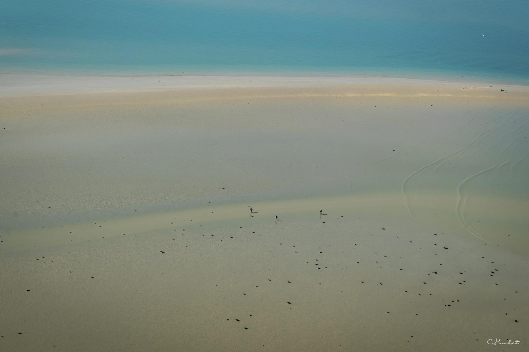 Immense étendue de sable blond parsemée de roches à marée basse avec quelques silhouettes humaines sous une mer turquoise.