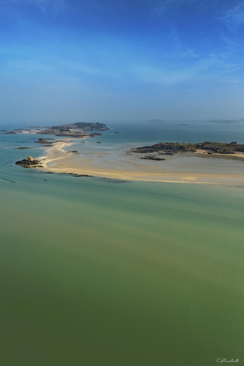 Vue aérienne de mer émeraude translucide, bancs de sable blanc et îlots rocheux bretons sous un ciel bleu dégagé.