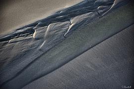 Textures de sable gris et ombres marquées d’un banc de sable lunaire en baie du Mont-Saint-Michel à marée basse.