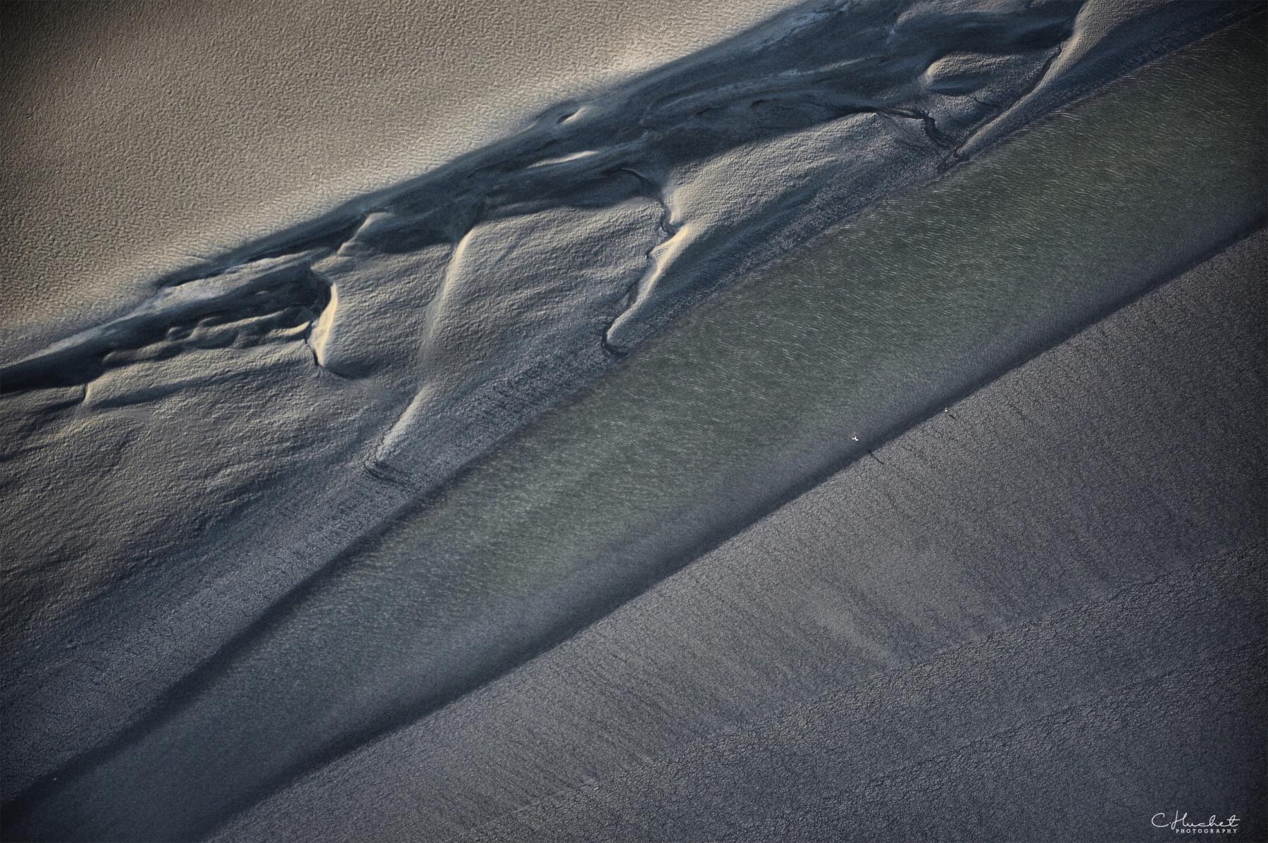 Textures de sable gris et ombres marquées d’un banc de sable lunaire en baie du Mont-Saint-Michel à marée basse.
