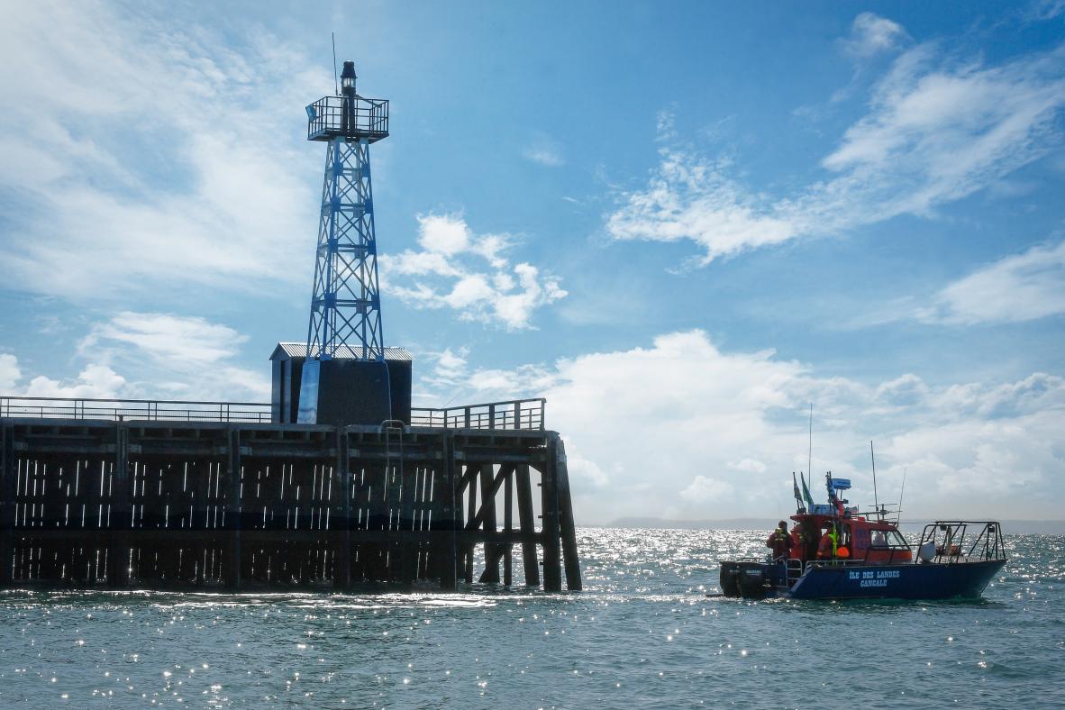 Vedette SNSM bleue quittant la jetée en bois de Cancale sous un ciel lumineux, structure de phare en arrière-plan.
