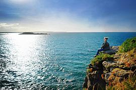 Silhouette contemplative assise au bord d’une falaise rocheuse devant une mer azur étincelante à Saint-Briac.