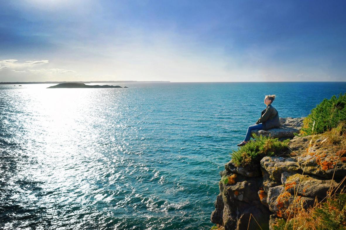 Silhouette contemplative assise au bord d’une falaise rocheuse devant une mer azur étincelante à Saint-Briac.