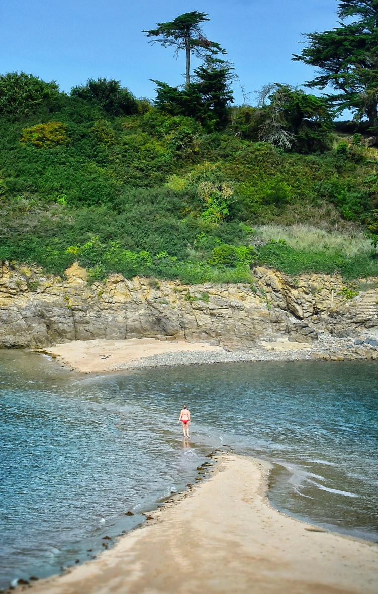 Femme en bikini rouge marchant sur un banc de sable submerge par l’eau turquoise devant une falaise fleurie.