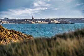 Silhouette de la cité corsaire de Saint-Malo vue par-delà l’eau bleue, cadrée depuis un premier plan de végétation sauvage.