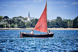 Voilier à voile rouge naviguant dans le Golfe du Morbihan face à l’île d’Arz sous une lumière estivale.