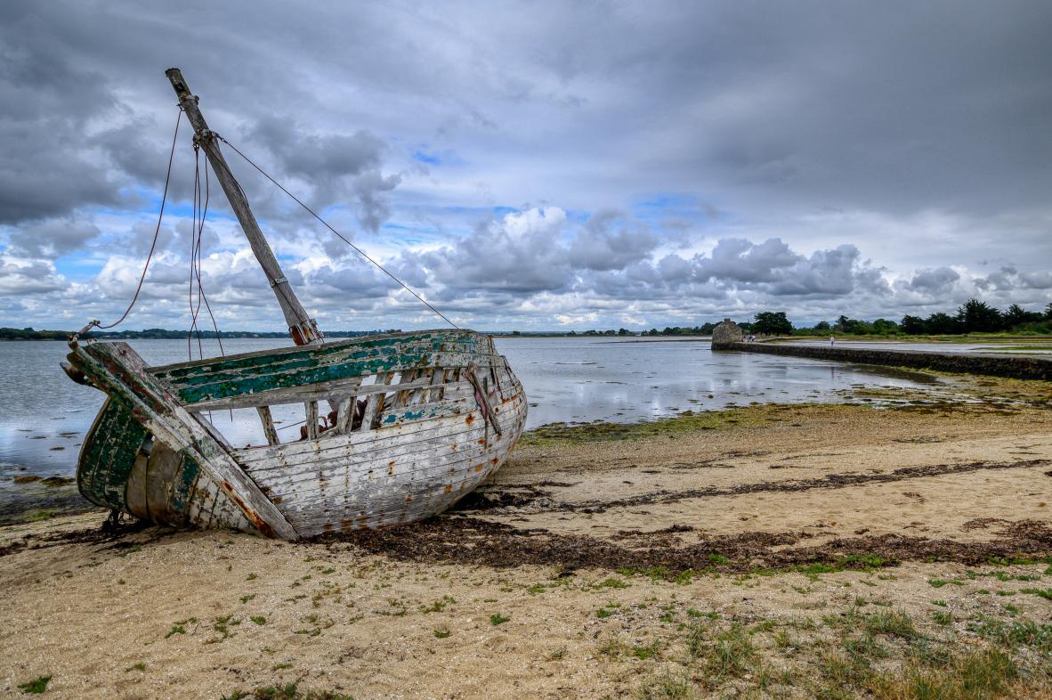Ancienne épave de bateau en bois vert et blanc échouée sur le sable devant le moulin de Berno sous un ciel nuageux.