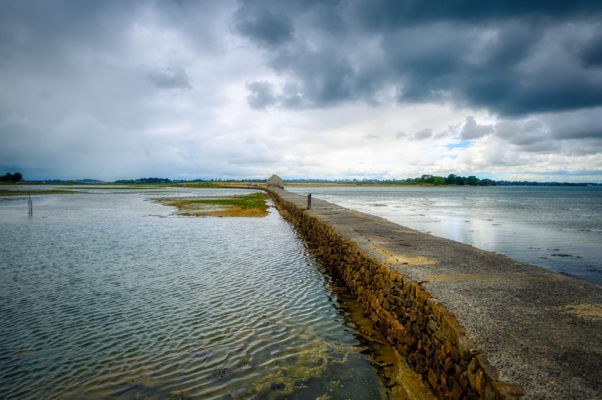 Digue en pierre vers le moulin de Berno sous un ciel chargé d’orage dans le Golfe du Morbihan en Bretagne.
