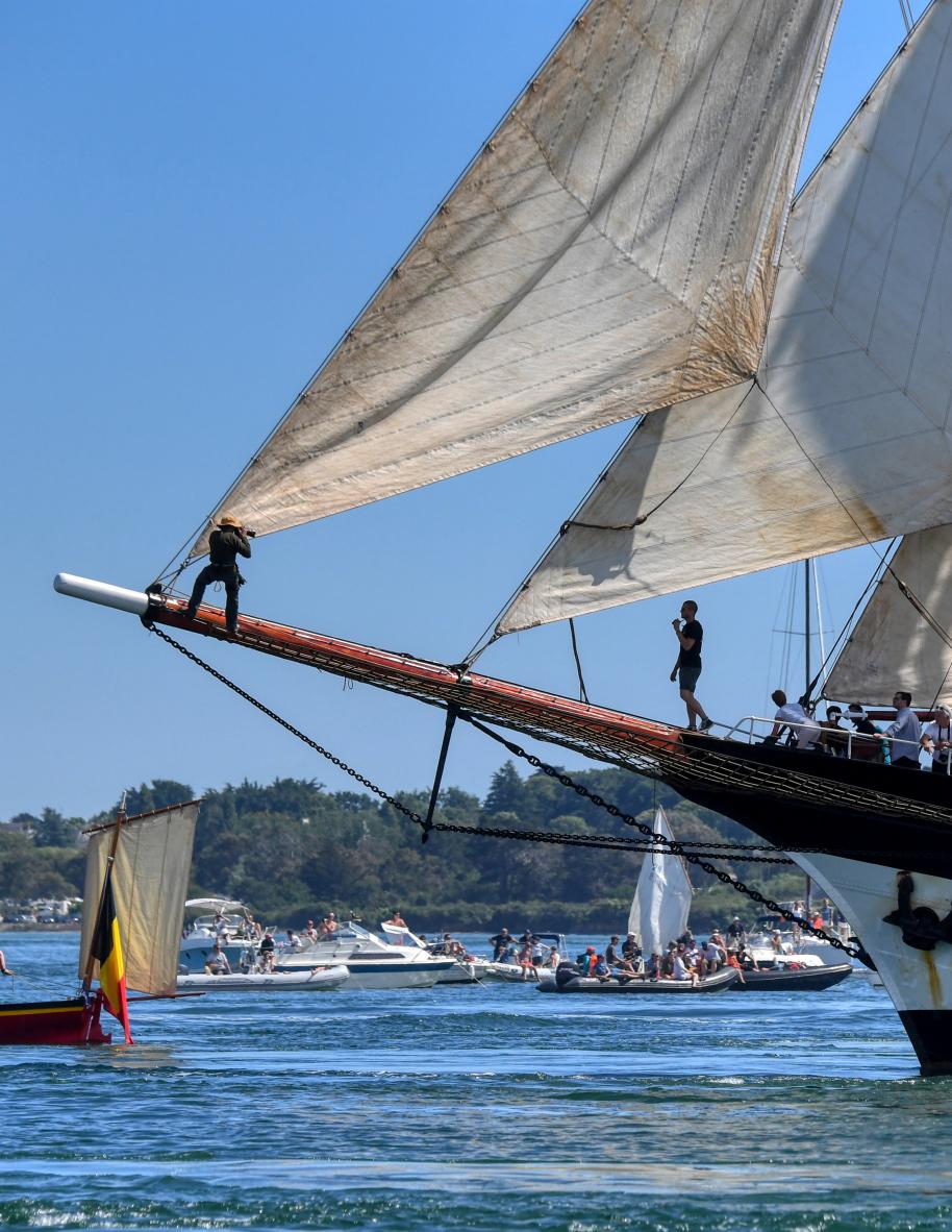Marin sur le beaupré d’un voilier traditionnel aux voiles blanches déployées dans le Golfe du Morbihan sous le soleil.