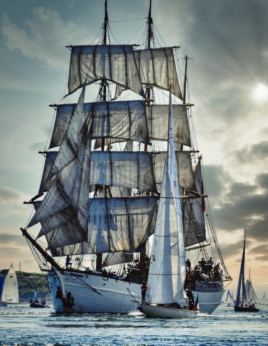 Grand trois-mâts aux voiles blanches déployées naviguant près d’un voilier moderne sur une mer calme en Bretagne.