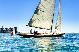Voilier classique 8mJ sous voiles blanches naviguant sur une mer bleue ensoleillée devant Port-Navalo en Bretagne.