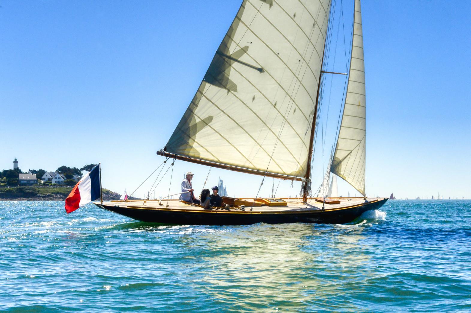 Voilier classique 8mJ sous voiles blanches naviguant sur une mer bleue ensoleillée devant Port-Navalo en Bretagne.