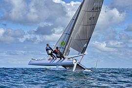 Catamaran blanc naviguant sur une coque en mer de Bretagne, deux équipiers en rappel sous un ciel nuageux.