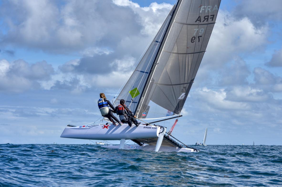 Catamaran blanc naviguant sur une coque en mer de Bretagne, deux équipiers en rappel sous un ciel nuageux.