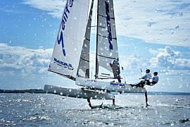 Catamaran Nacra sur foils volant au-dessus de l’eau avec des éclaboussures au premier plan sous un ciel nuageux.