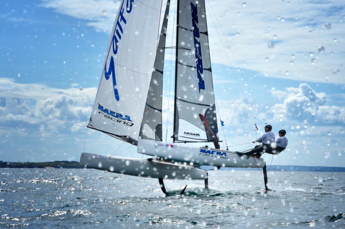 Catamaran Nacra sur foils volant au-dessus de l’eau avec des éclaboussures au premier plan sous un ciel nuageux.