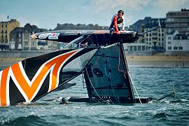Catamaran de régate renversé en mer à Saint-Malo, un marin sur la coque retournée sous un ciel lumineux.