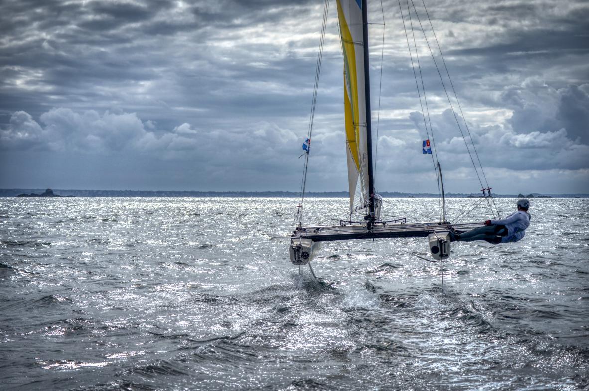 Catamaran à foils Flying Phantom naviguant à grande vitesse sur une mer scintillante sous un ciel nuageux à Saint-Malo.