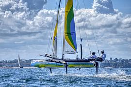 Catamaran Flying Phantom sur foils s’élevant au-dessus de l’eau devant la côte de Dinard sous un ciel nuageux.