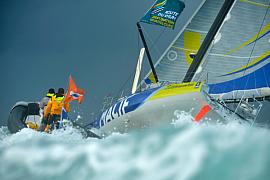 Imoca MACIF gîtant dans les vagues lors du départ de la Route du Rhum à Saint-Malo sous un ciel sombre.